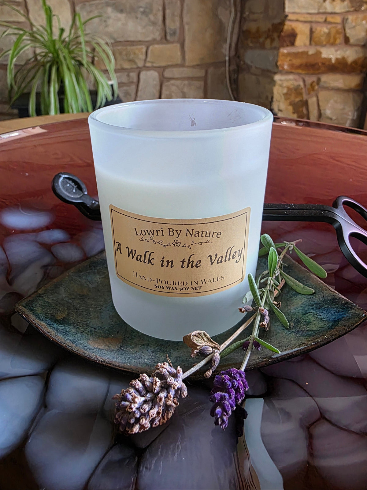 A frosted glass candle jar labeled 'Lowri By Nature A Walk in the Valley' sits on a rustic green ceramic plate with dried lavender sprigs, set on a reddish-brown stone surface with a stone wall background.