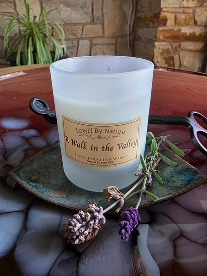 A frosted glass candle jar labeled 'Lowri By Nature A Walk in the Valley' sits on a rustic green ceramic plate with dried lavender sprigs, set on a reddish-brown stone surface with a stone wall background.