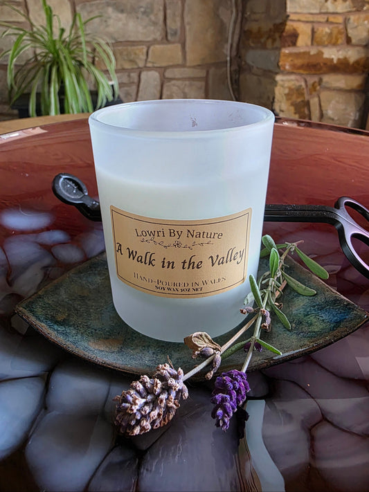 A frosted glass candle jar labeled 'Lowri By Nature A Walk in the Valley' sits on a rustic green ceramic plate with dried lavender sprigs, set on a reddish-brown stone surface with a stone wall background.
