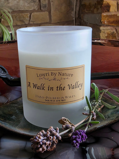 A frosted glass candle jar labeled 'Lowri By Nature A Walk in the Valley' sits on a rustic tray with dried lavender sprigs, set against a warm stone and wood background.