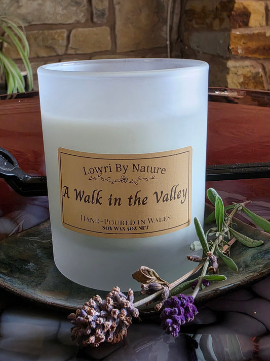 A frosted glass candle jar labeled 'Lowri By Nature A Walk in the Valley' sits on a rustic tray with dried lavender sprigs, set against a warm stone and wood background.