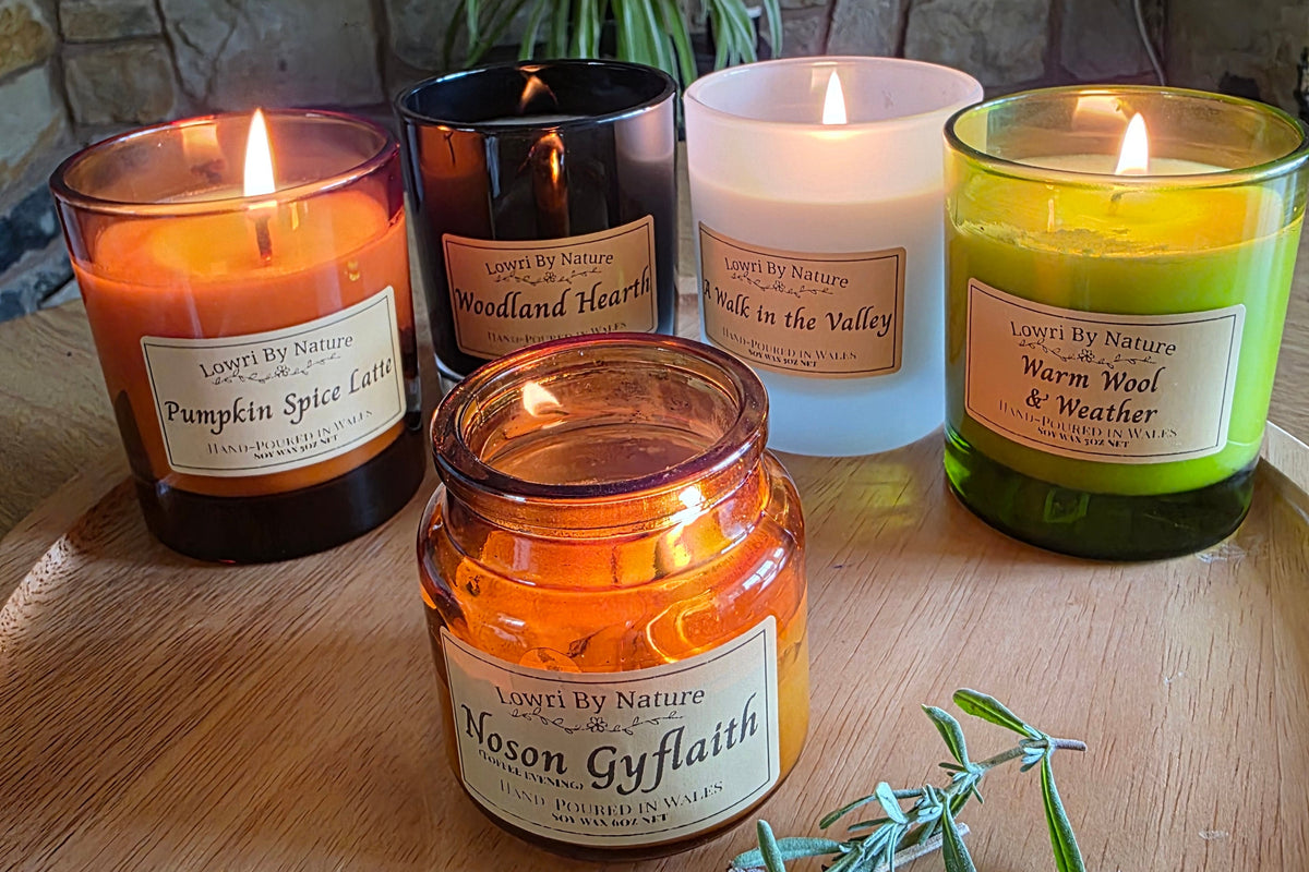 Six scented candles in glass jars arranged on a wooden tray in front of a stone fireplace. The candles include amber, dark brown, white, and green glass jars, each with a lit flame and a paper label.