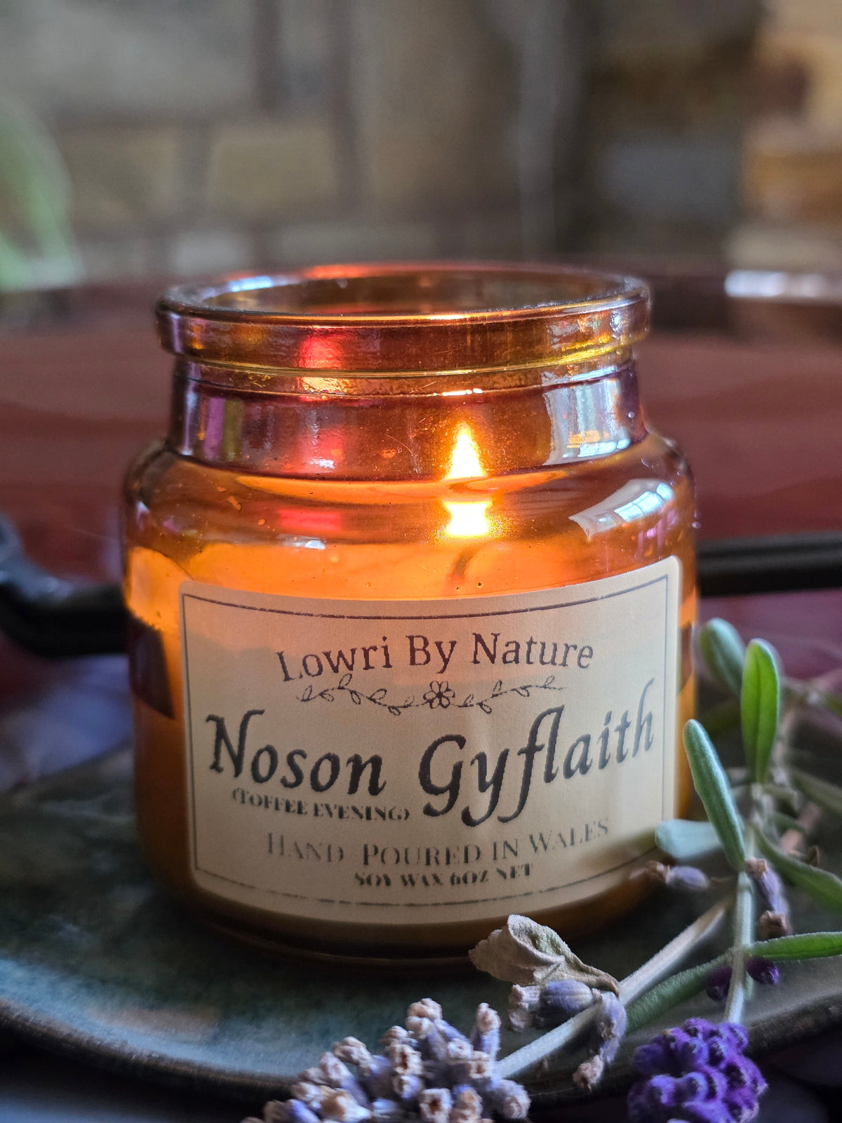 A lit amber glass jar candle with a cream label reading 'Lowri By Nature Noson Gyflaith (Toffee Evening)', styled on a dark plate with lavender sprigs in a warm indoor setting.