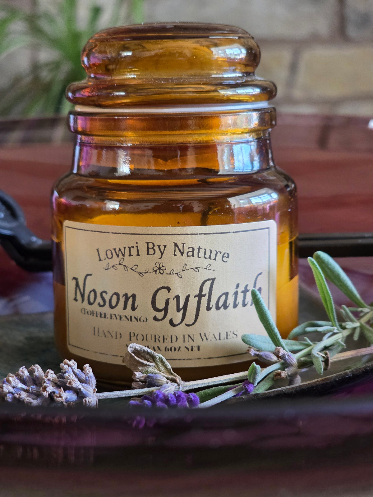 An amber glass jar candle with a cream label reading 'Lowri By Nature Noson Gyflaith' and 'Hand Poured in Wales', styled on a dark tray with lavender sprigs and greenery.