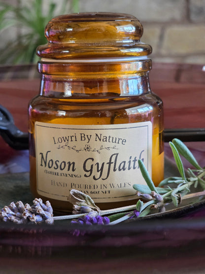 An amber glass jar candle with a cream label reading 'Lowri By Nature Noson Gyflaith' and 'Hand Poured in Wales', styled on a dark tray with lavender sprigs and greenery.