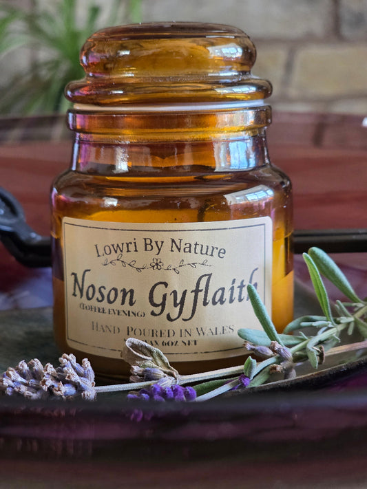 An amber glass jar candle with a cream label reading 'Lowri By Nature Noson Gyflaith' and 'Hand Poured in Wales', styled on a dark tray with lavender sprigs and greenery.