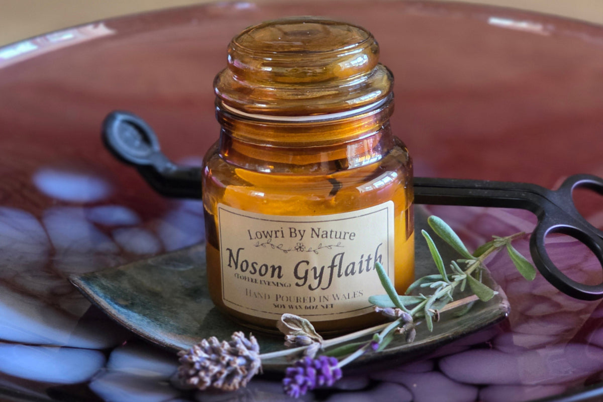 A small amber glass candle jar labeled Lowri By Nature Noson Gyflait' sits on a dark ceramic plate with purple marbled patterns, styled with dried lavender sprigs on a wooden table.