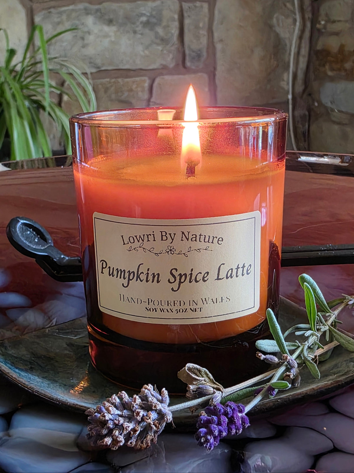 A lit candle in a clear glass jar labeled 'Lowri By Nature Pumpkin Spice Latte,' placed on a tray with lavender sprigs in front of a stone wall background.
