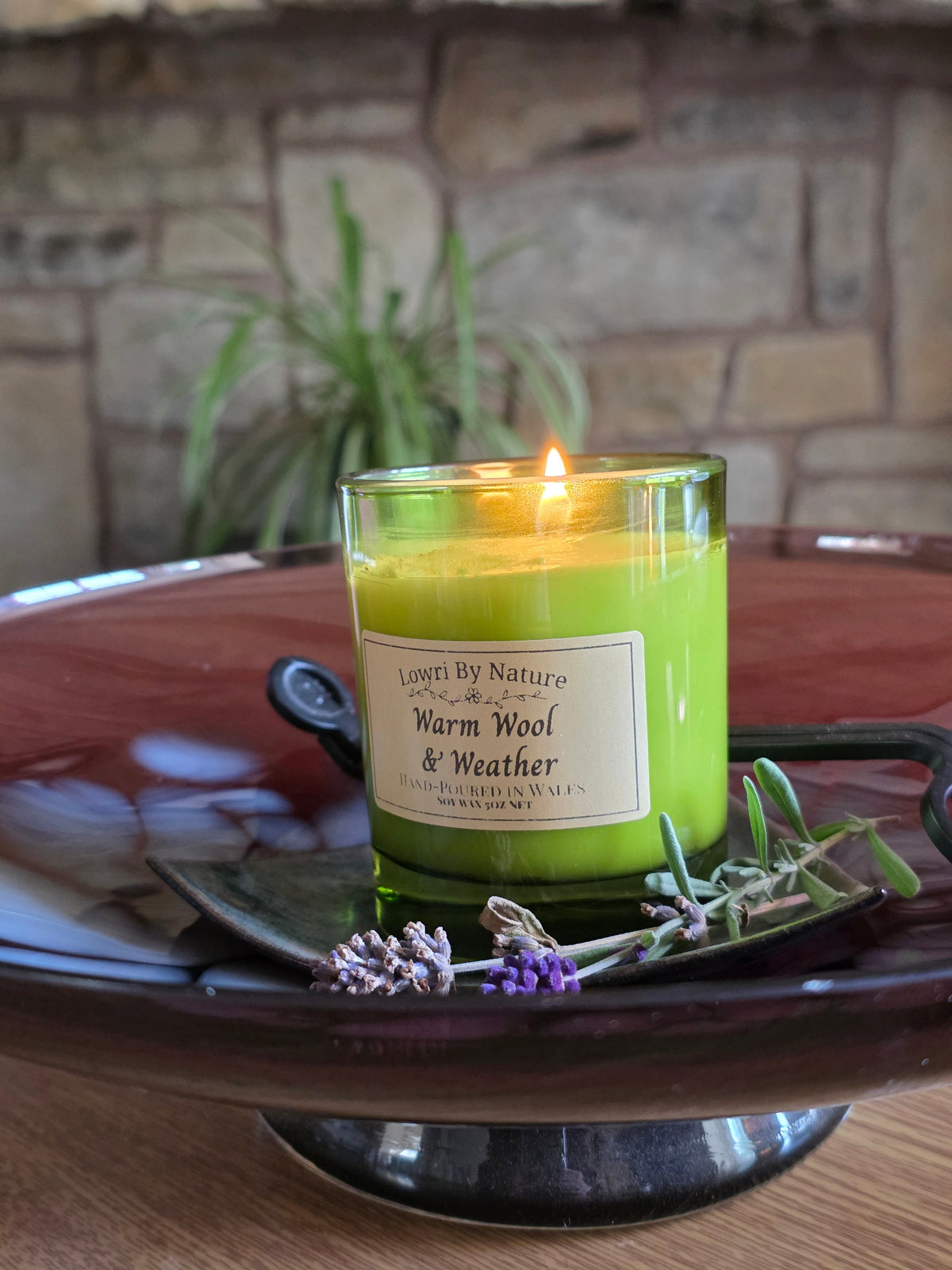 A lit candle in a clear glass jar with bright yellow-green wax sits on a dark ceramic tray, surrounded by lavender sprigs. The label reads 'Lower By Nature Warm Wool & Weather'.