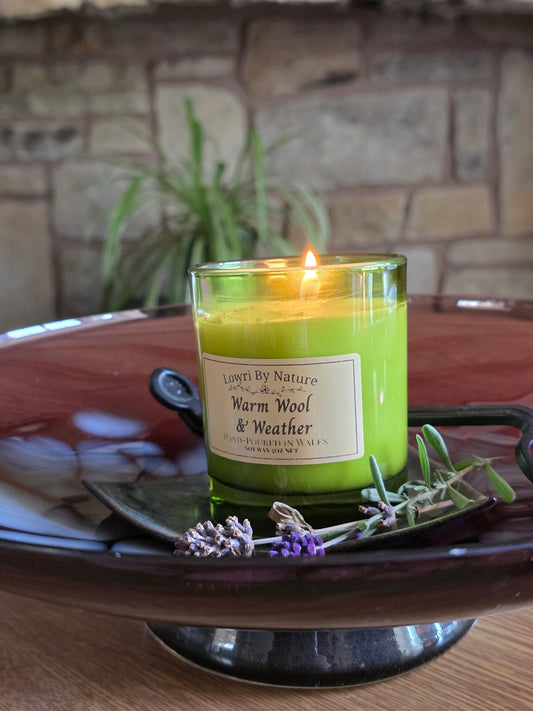 A lit candle in a clear glass jar with bright yellow-green wax sits on a dark ceramic tray, surrounded by lavender sprigs. The label reads 'Lower By Nature Warm Wool & Weather'.