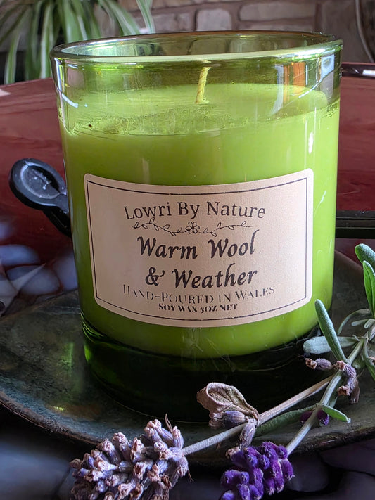 A green soy wax candle in a clear glass jar with a cream label reading 'Lowri By Nature Warm Wool & Weather'. The candle is styled on a dark plate with lavender sprigs and a dark spoon against a rustic background.
