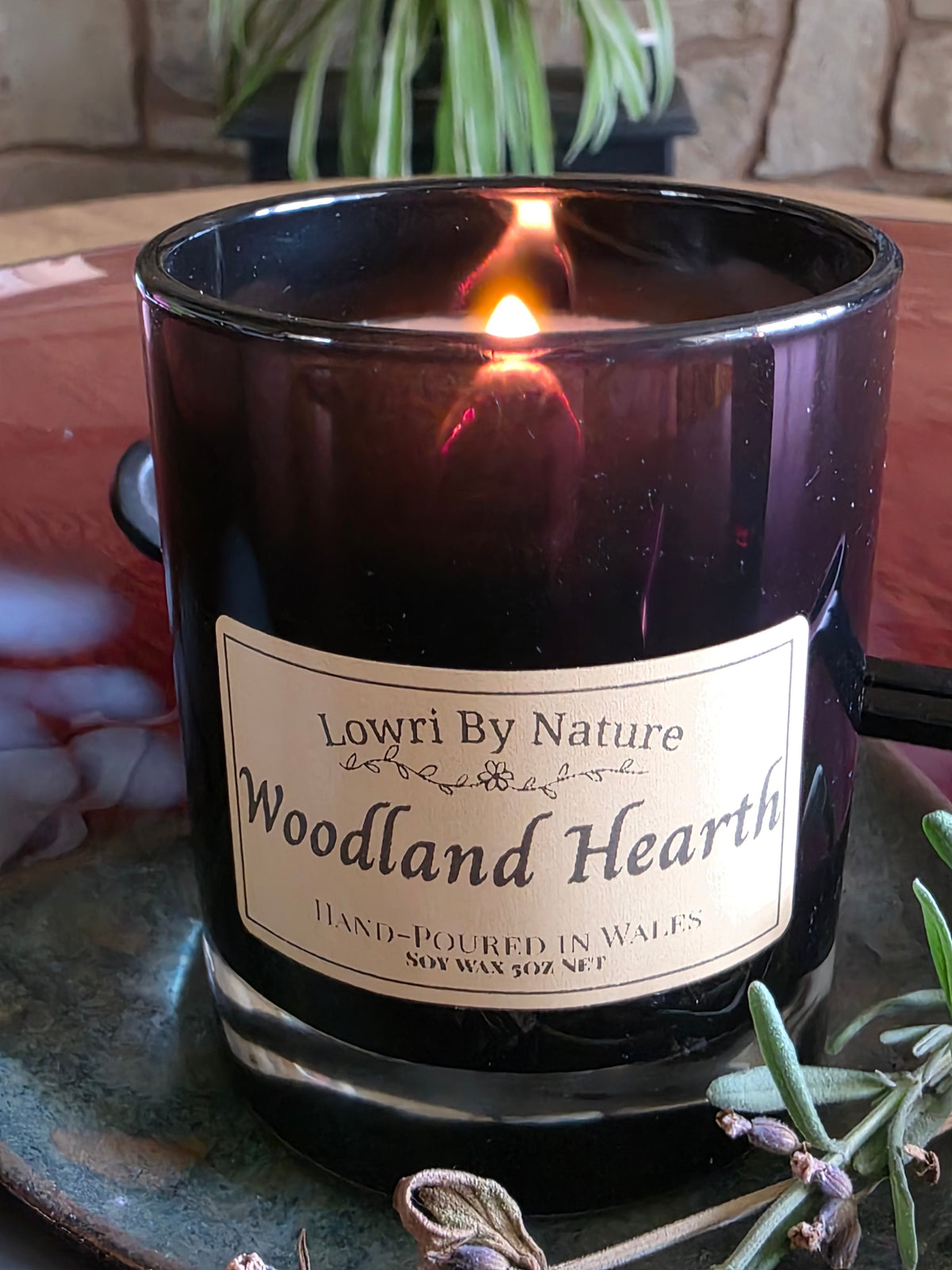 A lit candle in a black glass jar with a sand label reading 'Lowri By Nature Woodland Hearth,' shown on a rustic table with a plant and greenery in the background.