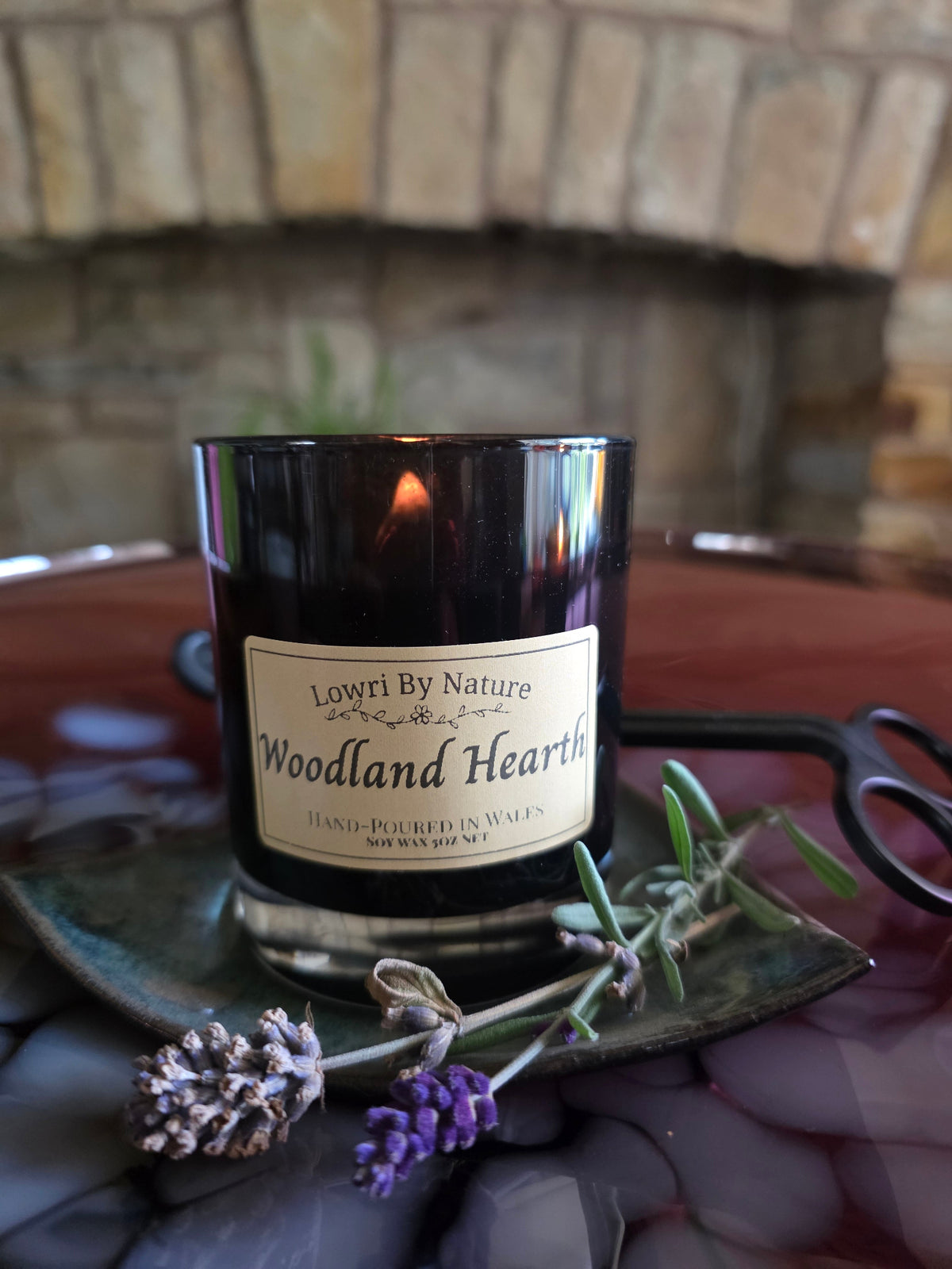 A black glass jar candle labeled 'Lowri By Nature Woodland Hearth' sits on a reflective dark tray with lavender sprigs and a black metal tool, set against a blurred stone fireplace background.